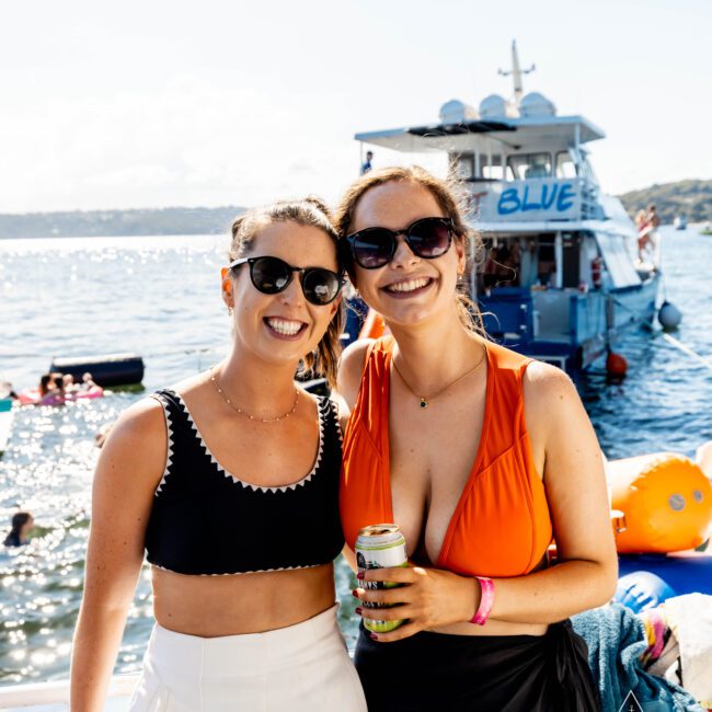 Two smiling women in sunglasses stand close together at a sunny boat party. One wears a black crop top and white skirt; the other, an orange swimsuit. They are holding drinks, with a boat named “BLUE” from The Yacht Social Club Sydney Boat Hire and floating inflatables visible in the background.