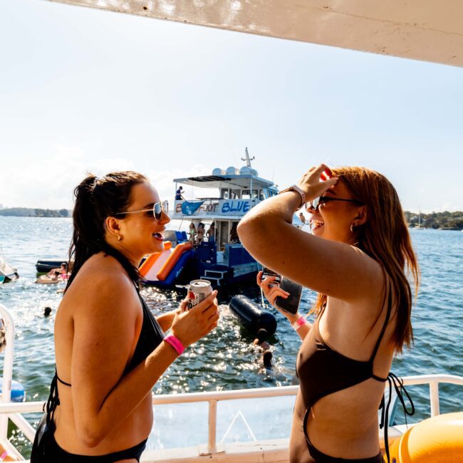 Two women in swimsuits stand on a boat, laughing and talking by the railing. The water is calm with several boats and colorful recreational inflatables in the background. A sunny day with clear skies provides a joyful and relaxed atmosphere, perfect for Boat Parties Sydney with The Yacht Social Club.