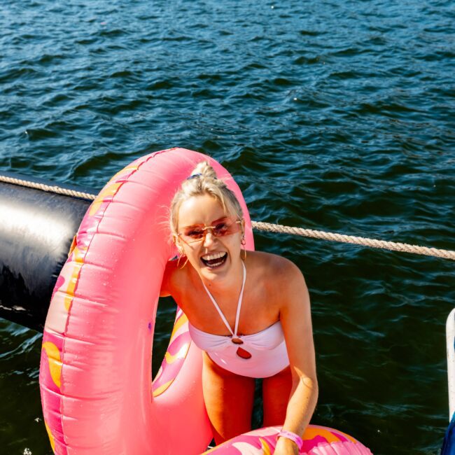 A smiling woman in a white swimsuit stands in the water, holding a large pink inflatable pool ring. She has light-colored hair tied up and is wearing pink sunglasses. The background features calm water and a distant, tree-lined shore—perfect for enjoying The Yacht Social Club Event Boat Charters.