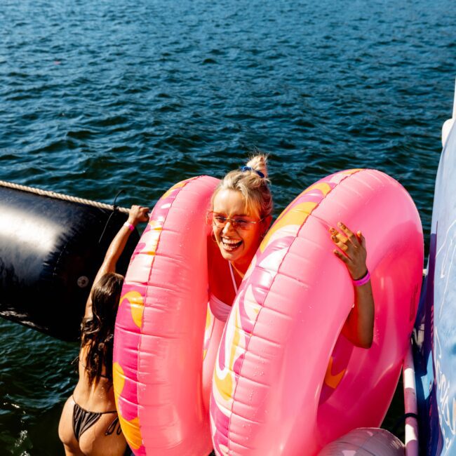 A person with light hair in a bun, wearing sunglasses, is smiling and standing on a dock with two pink inflatable rings around them. Another person in a swimsuit is partially visible, climbing onto the dock from the water. The backdrop evokes vibes of Luxury Yacht Rentals Sydney with its scenic lake and trees.