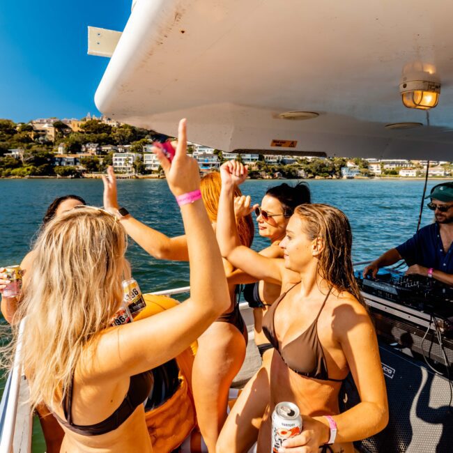 A lively group in swimwear dances and socializes on a boat under sunny skies. A DJ plays music as others enjoy the atmosphere. The Yacht Social Club organizes these luxurious gatherings, with the scenic shoreline of Sydney in the distance. Bright and festive scene!