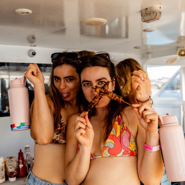 Two women in swimwear are posing for a playful photo on a boat. One holds a thermos and sticks out her tongue, while the other holds a skewer of food to her mouth and smiles. Bottles of condiments are visible in the background aboard The Yacht Social Club Sydney Boat Hire.