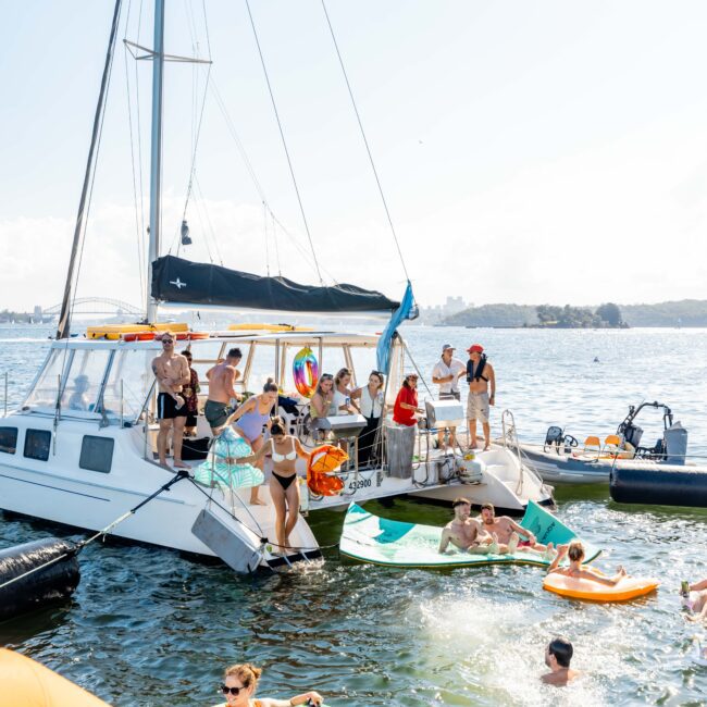 A group of people are enjoying a sunny day on the water at The Yacht Social Club Event Boat Charters. Some are on a sailboat, while others float in the water on inflatables. People are swimming and socializing, surrounded by boats and watercrafts. The scene is lively with clear, blue water and a bright sky.