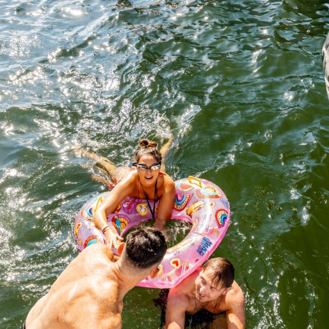 People enjoying a day in the water, with a man helping a woman in a colorful inflatable ring climb a boat ladder. Other swimmers are visible in the background, with sunlight reflecting off the water. The words "The Yacht Social Club Event Boat Charters" are visible in the bottom right corner.