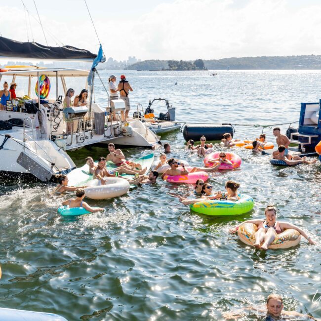 A vibrant scene of people enjoying a sunny day on the water. Several individuals are floating on colorful inflatables near anchored boats from The Yacht Social Club Event Boat Charters. Some are sunbathing on the boats, and an inflatable slide is visible. The distant shoreline and cityscape are in the background.