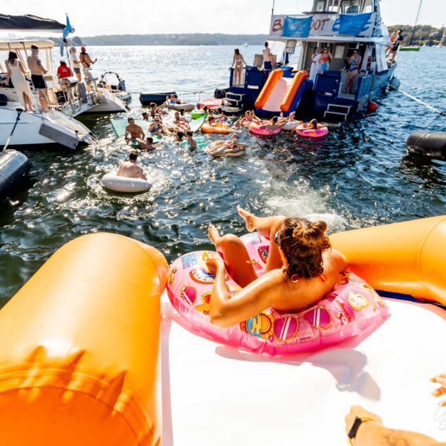 People are enjoying a sunny day on the water with several boats and floats around. A person slides down a white and orange inflatable slide while sitting in a pink donut-shaped float. Others swim or float nearby with vibrant pool floats. The Yacht Social Club Event Boat Charters offer unforgettable experiences.