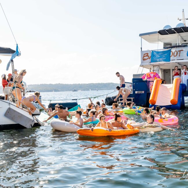 The Yacht Social Club is hosting a lively boat party in Sydney with numerous people on inflatables enjoying the water. Several boats, including a large one with a slide, are docked together. Sunny weather and clear skies enhance the festive atmosphere, with the horizon visible in the background.