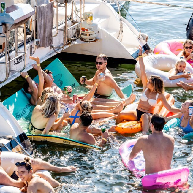 A group of people enjoying a sunny day on the water, gathered around boats and floating on various inflatables. Some are holding drinks, chatting, and laughing. In the background, boats from The Yacht Social Club are moored together, creating a lively and festive atmosphere.