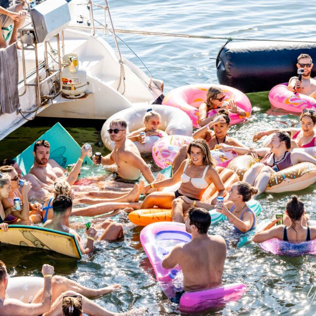 A group of people enjoying a sunny day on the water, lounging on various colorful inflatable floats near a boat. Many are holding drinks and wearing swimwear, with some standing in the water and others relaxing on the floats. The scene is lively and festive, perfect for Sydney Harbour Boat Hire with The Yacht Social Club.