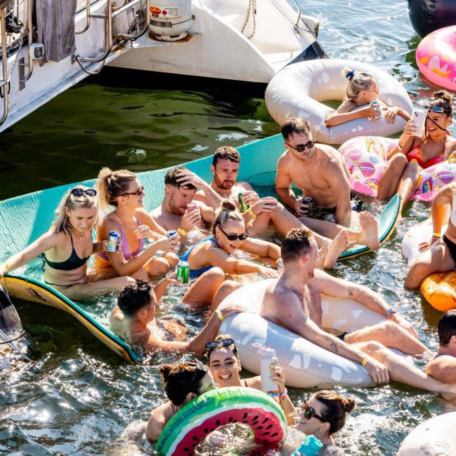 A group of people enjoying a sunny day on a river. They are on various inflatable pool floats and a floating mat, holding drinks. Boats from The Yacht Social Club Sydney Boat Hire are docked nearby, and the atmosphere is lively and festive with people chatting and relaxing in swimwear.