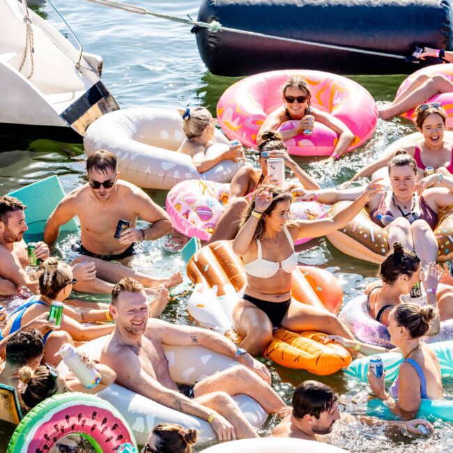 A group of people enjoying a sunny day in the water, sitting on colorful inflatable pool floats including a donut, watermelon slice, and pizza slice. They are smiling, chatting, and holding drinks, with a boat from Sydney Harbour Boat Hire The Yacht Social Club partially visible in the background.