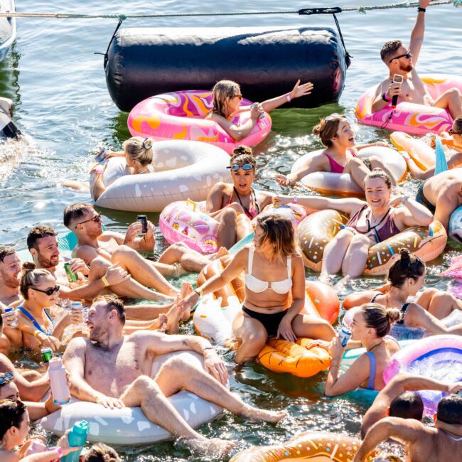 A lively group of people enjoys a sunny day floating on inflatable pool floats in the water. They are smiling, laughing, and socializing. Nearby, a boat from The Yacht Social Club is anchored, and various colorful floats, including a flamingo and a doughnut, add to the festive scene.