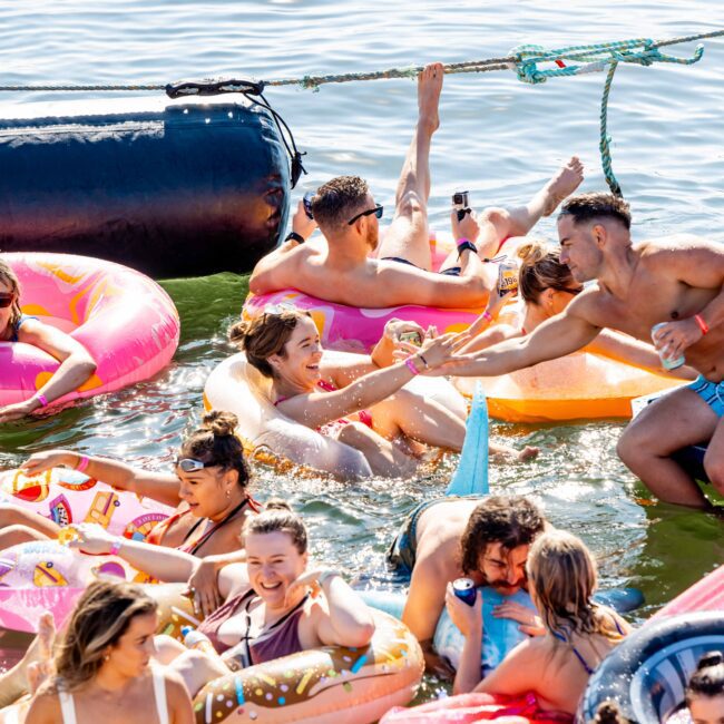 A group enjoys a sunny day at a lake, floating on colorful inflatables. Some are holding drinks, and one person in blue trunks leans from a dock to offer a drink to another in the water. They are all smiling and having a fun time, reminiscent of events hosted by The Yacht Social Club Event Boat Charters.