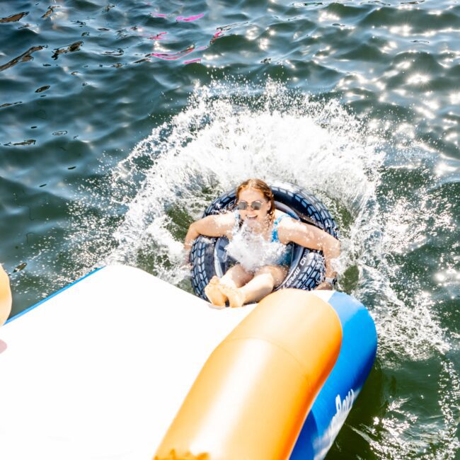 A young girl with sunglasses splashes into the water from an inflatable slide, landing in an inner tube. Several other inflatables and people are visible in the background, enjoying a sunny day on Sydney Harbour Boat Hire The Yacht Social Club.