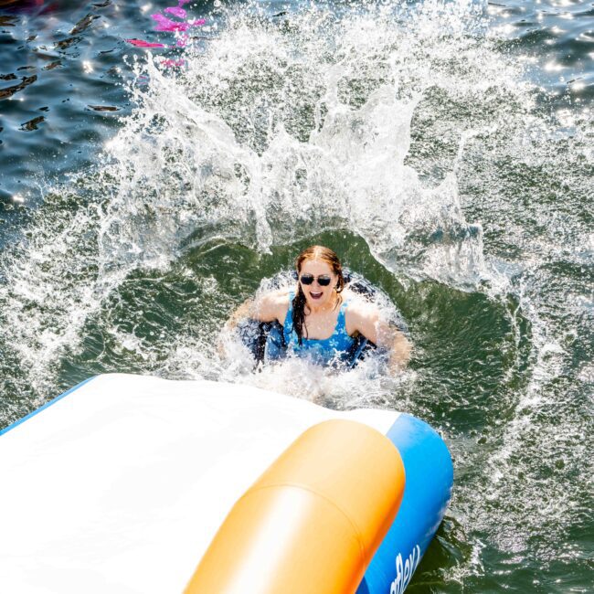 A person with sunglasses is splashing into the water from an inflatable slide. Other people float on colorful inflatables in the background. The sunlight casts a bright reflection on the water's surface. Text at the bottom right corner reads "The Yacht Social Club.