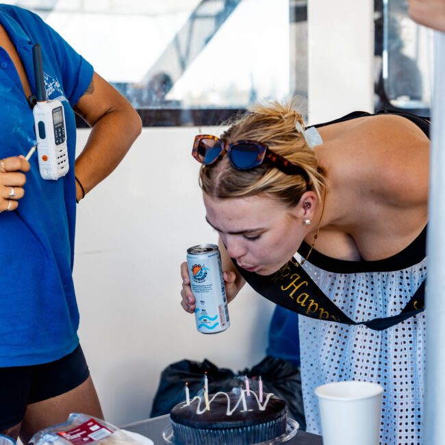 A woman with a sash leans over to blow out candles on a birthday cake. She holds a drink can and wears sunglasses on her head. Another person in a blue shirt stands nearby holding a radio. They are on a boat, evidenced by The Yacht Social Club logo, enjoying one of their Boat Rental and Parties Sydney events.