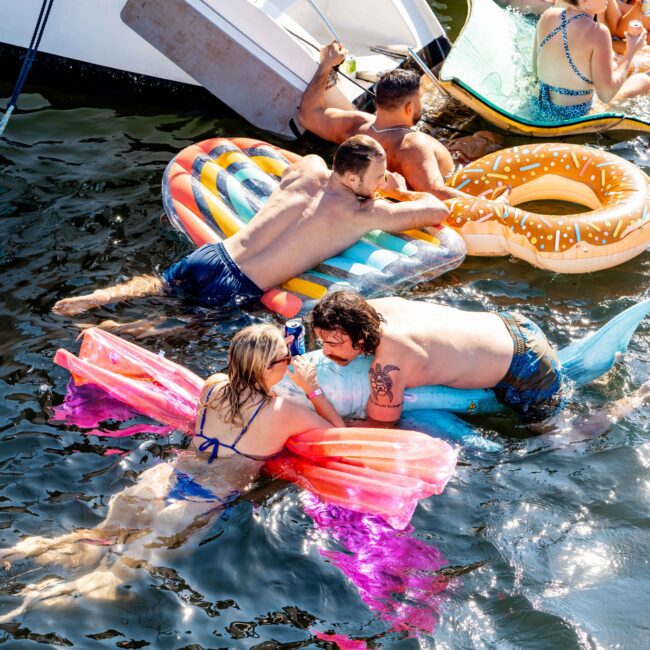 A group of people enjoys a sunny day on inflatables in the water near a docked boat from The Yacht Social Club Sydney Boat Hire. They are relaxing and chatting, with some on colorful floaties. A woman in a blue bikini is on a pink floatie, conversing with a man on a blue floatie.