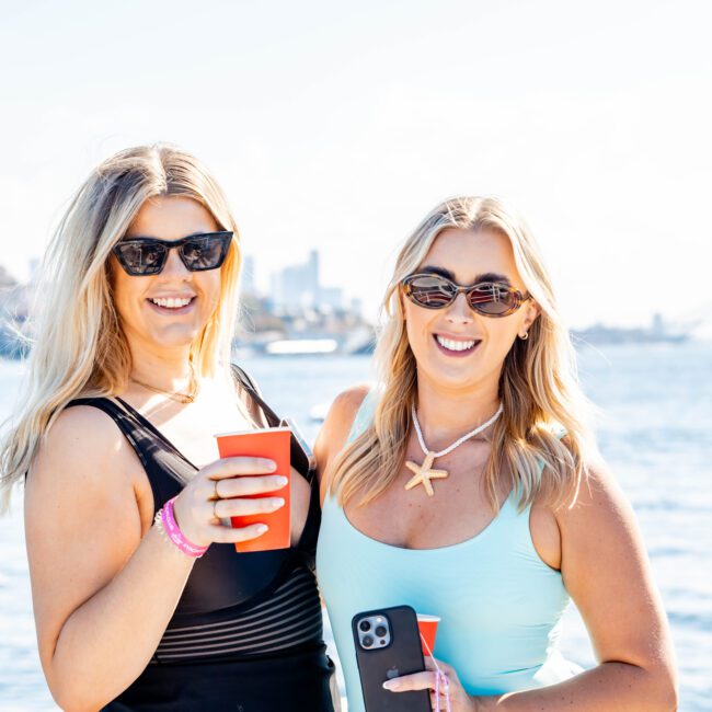 Two smiling women stand side by side by the water, holding red cups and wearing sunglasses. One wears a black dress, and the other a light blue dress with a starfish necklace. Both have smartphones on wristbands, with the stunning city skyline in the background, enjoying The Yacht Social Club Event Boat Charters.