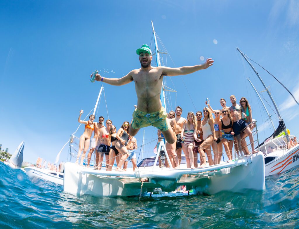 A group of people aboard a sailboat is enjoying a sunny day on the water. In the foreground, a man wearing swim trunks and a hat is captured mid-jump off the boat into the water. The group behind him from Boat Parties Sydney The Yacht Social Club is cheering and holding drinks.