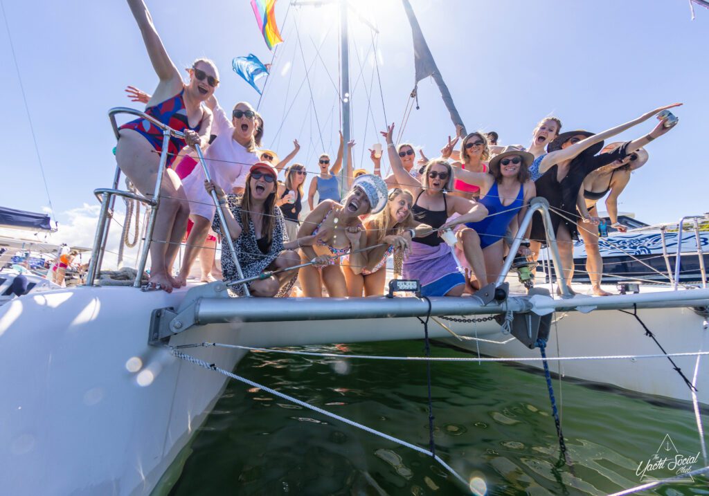 A group of people in colorful swimwear are gathered at the edge of a boat, smiling and making celebratory gestures. They are enjoying a sunny day with The Yacht Social Club Sydney Boat Hire. Some hold drinks and one person wears a snorkel mask. The water and other boats are visible in the background.