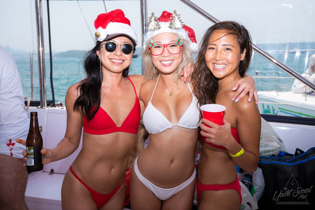 Three women in bikinis enjoying a festive boat party by The Yacht Social Club Sydney Boat Hire. Two women are wearing red bikinis with Santa hats and sunglasses; the woman in the middle is in a white bikini with decorative holiday glasses. They are smiling and holding drinks, with the sea visible in the background.