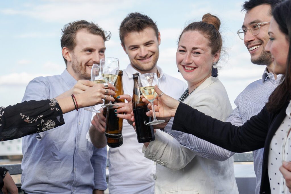 A group of five people is standing together, smiling, and holding beverages, including beer bottles and glasses of wine. They are toasting and clinking their drinks against a backdrop of a clear sky, enjoying the celebration at The Yacht Social Club Event Boat Charters. Everyone appears to be in a celebratory mood.