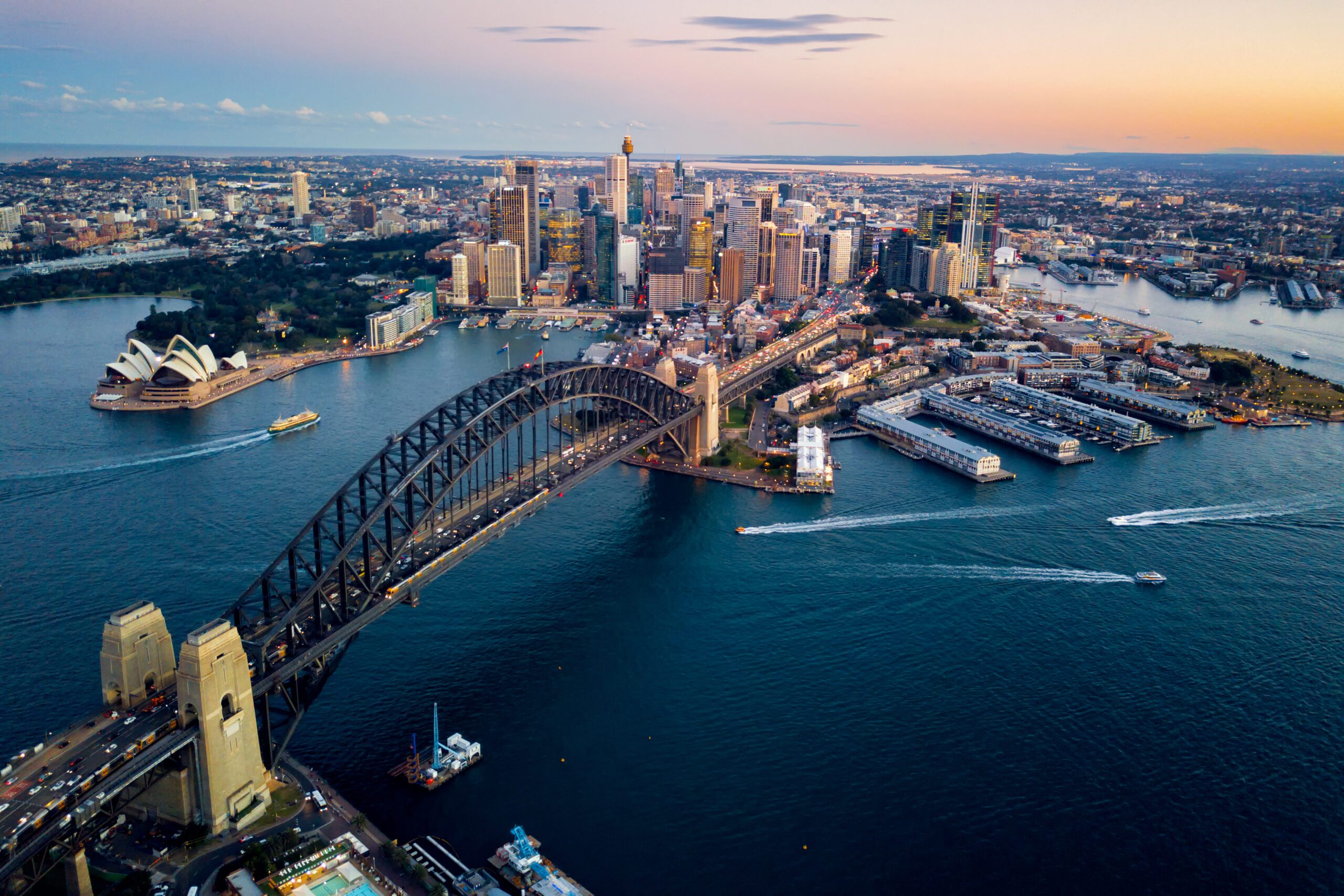 Aerial view of Sydney, Australia at sunset, featuring the Sydney Harbour Bridge spanning over the water, the Sydney Opera House on the left, and the city's skyline in the background. Boats from The Yacht Social Club Sydney Boat Hire are visible in the harbor, and the sky is painted in warm hues.