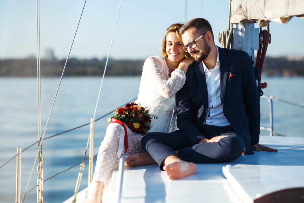 A happy couple sits together on the deck of a sailboat, with a calm body of water in the background. The woman, wearing a white dress, holds a bouquet and rests her head on the man's shoulder. The man is dressed in a dark suit and glasses, smiling contentedly at The Yacht Social Club Event Boat Charters.