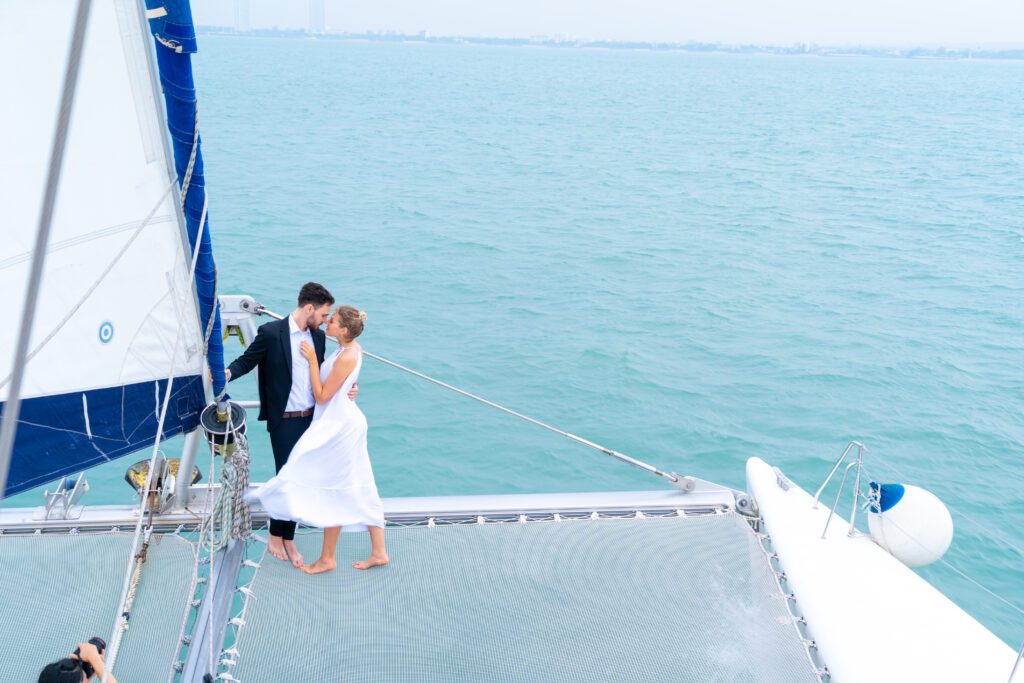 A bride and groom share an intimate moment on the deck of a sailboat. The wind ruffles the bride's white dress, and the groom is in a black suit. They are surrounded by calm, expansive blue water under a light, cloudy sky, enjoying their special day with Luxury Yacht Rentals Sydney.