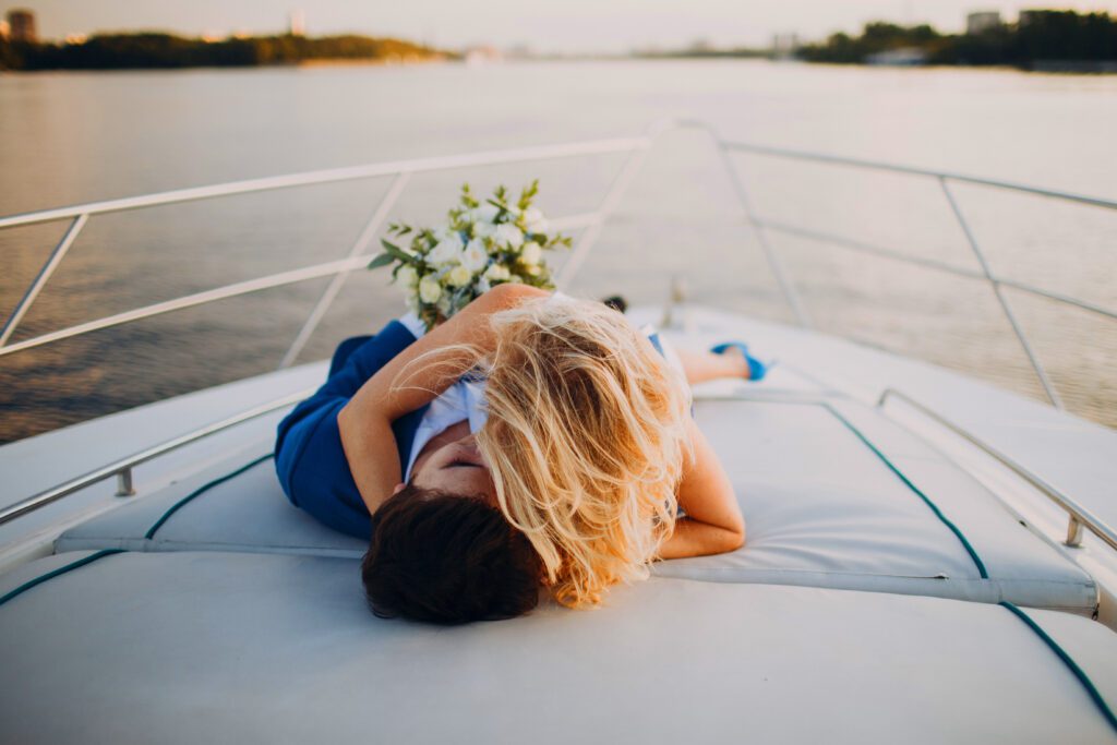 A couple is lying down and embracing on the deck of a luxury yacht at sunset. They are surrounded by calm waters, with a scenic view of the distant shoreline. A bouquet of flowers lies beside them, adding a romantic touch to the peaceful setting—a perfect moment courtesy of Luxury Yacht Rentals Sydney.