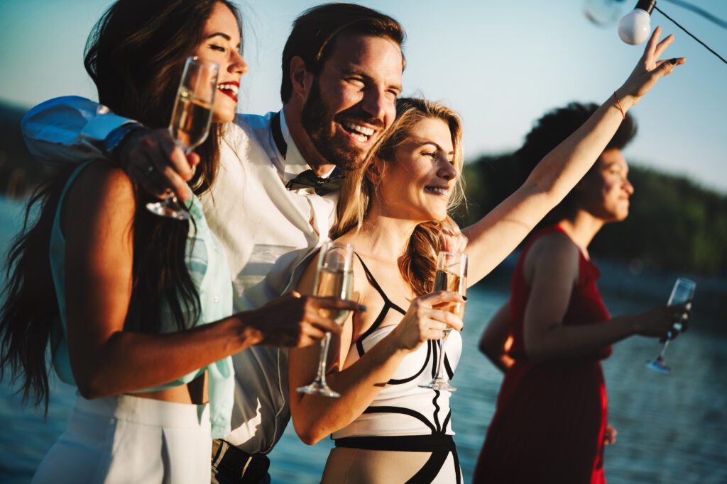 Four people are enjoying an outdoor celebration by a body of water, as part of The Yacht Social Club Sydney Boat Hire. Three are in the foreground, smiling and holding glasses of champagne, with one person raising their arm joyously. The fourth person is in the background, gazing away. The mood is festive and cheerful.