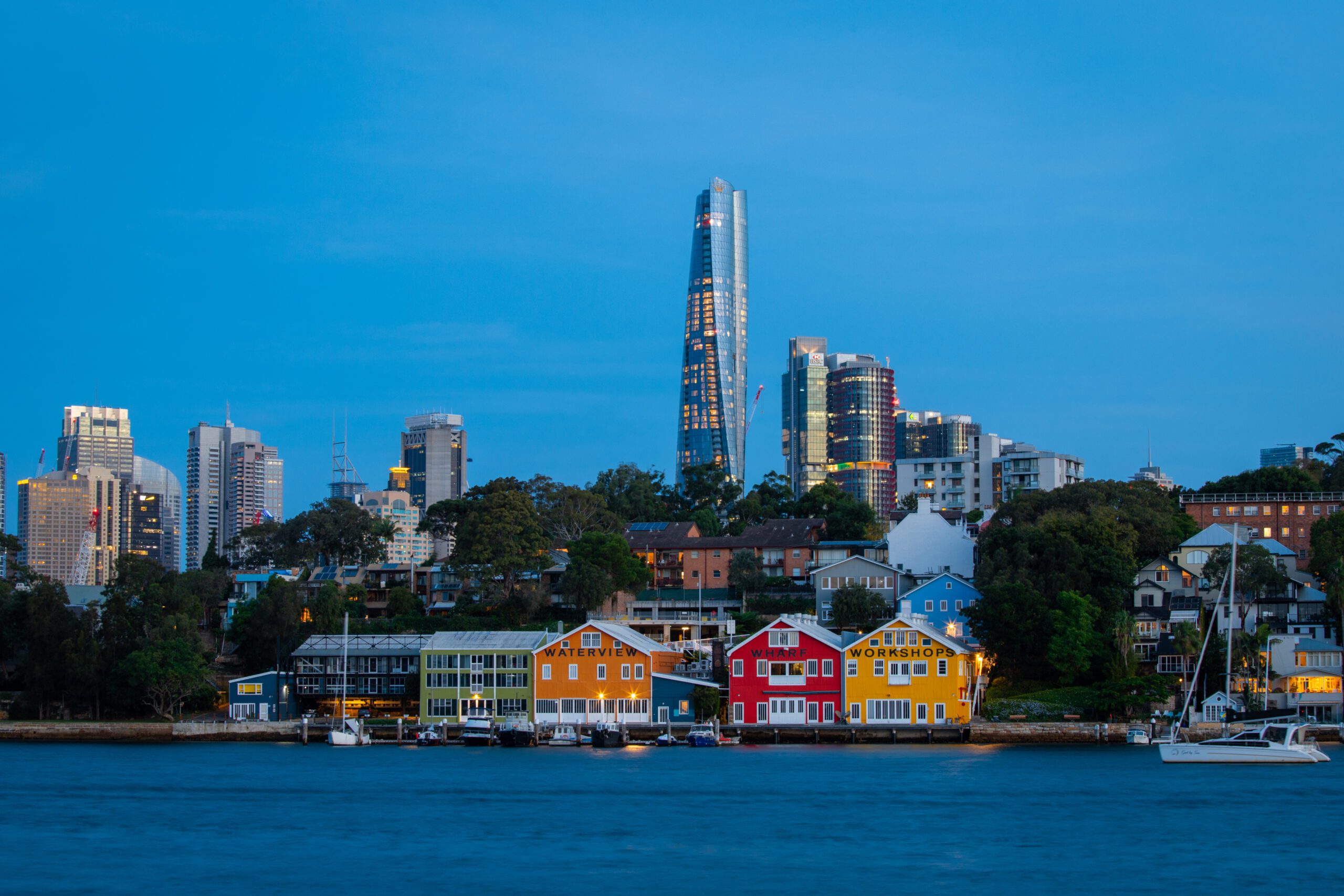 A vibrant waterfront scene at dusk features colorful buildings and boats docked by the water, hinting at exciting boat rental adventures with The Yacht Social Club. In the background, a modern city skyline with various skyscrapers rises against a clear blue sky, while trees and smaller buildings are scattered throughout the area.