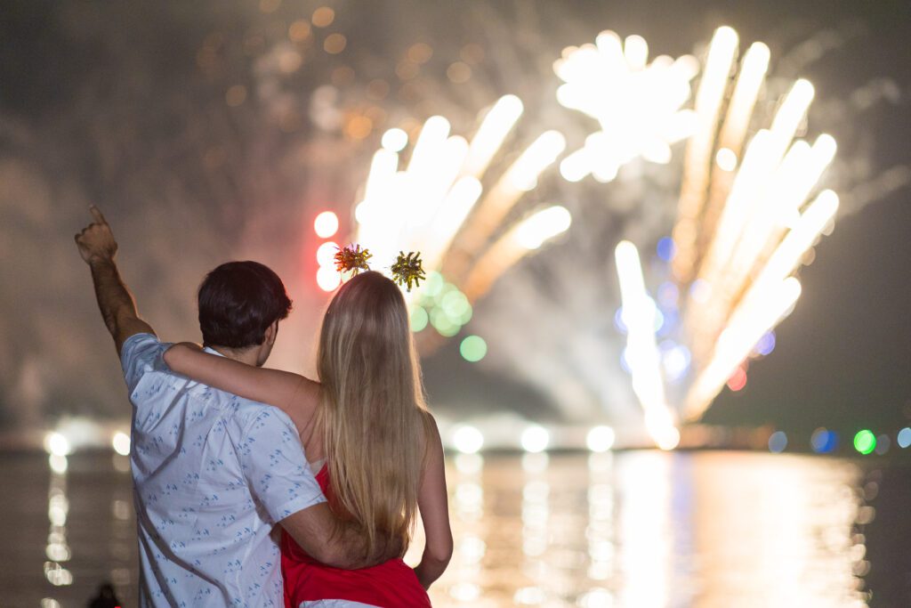 A couple stands by a body of water at night, watching a vibrant fireworks display. The man has his arm around the woman, pointing towards the sky. The fireworks illuminate the night, casting reflections on the water's surface and enhancing their experience with Sydney Harbour Boat Hire's services from The Yacht Social Club.