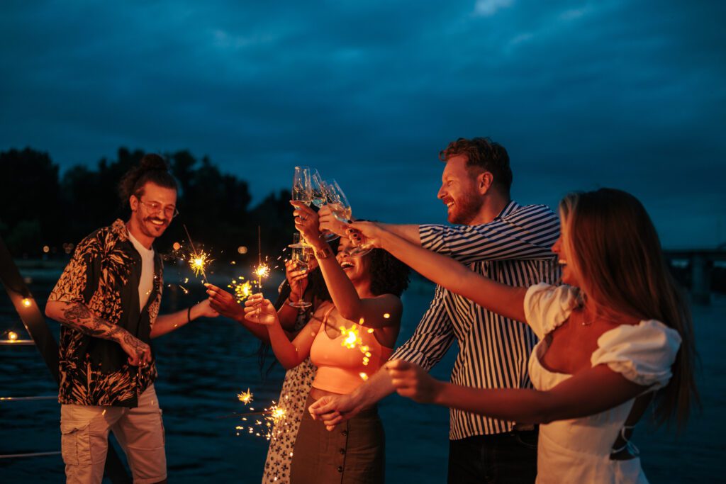 A group of five people celebrate outdoors at dusk, holding sparklers and raising glasses in a toast. They smile and laugh against a backdrop of darkening sky and water, indicating a joyful gathering or party by a lake or river—perhaps part of the lively boat parties with The Yacht Social Club in Sydney Harbour.