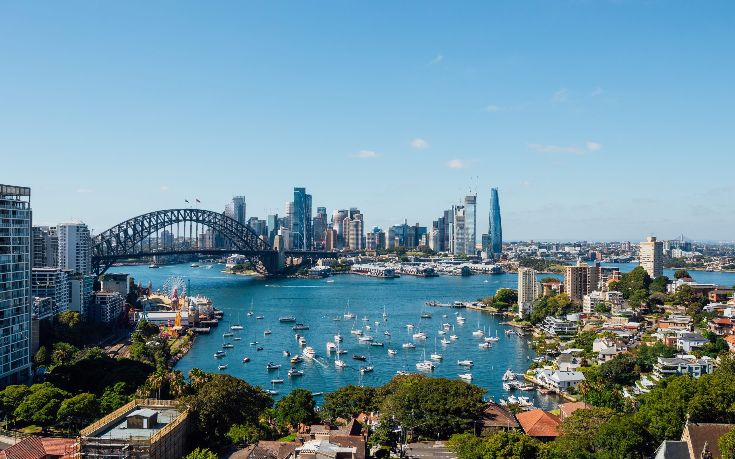 A scenic view of Sydney Harbour features the iconic Sydney Harbour Bridge, numerous boats from The Yacht Social Club scattered on the vibrant water, and the city's high-rise buildings in the background under a sunny, clear blue sky. The coastline is dotted with greenery and residential houses.