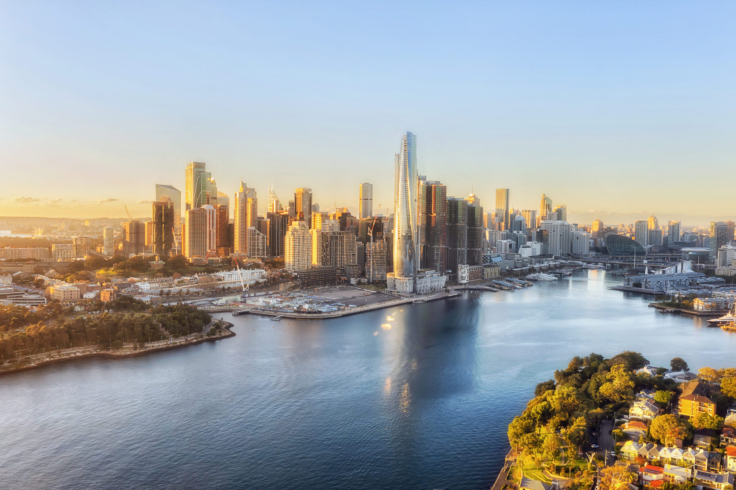 Aerial view of a cityscape at sunset, featuring a river flanked by a mix of tall skyscrapers and smaller buildings. The skyline reflects in the calm river, with a park visible in the foreground. The scene feels elegant, hinting at luxury yacht rentals available nearby. The sky is clear, bathed in warm golden light.