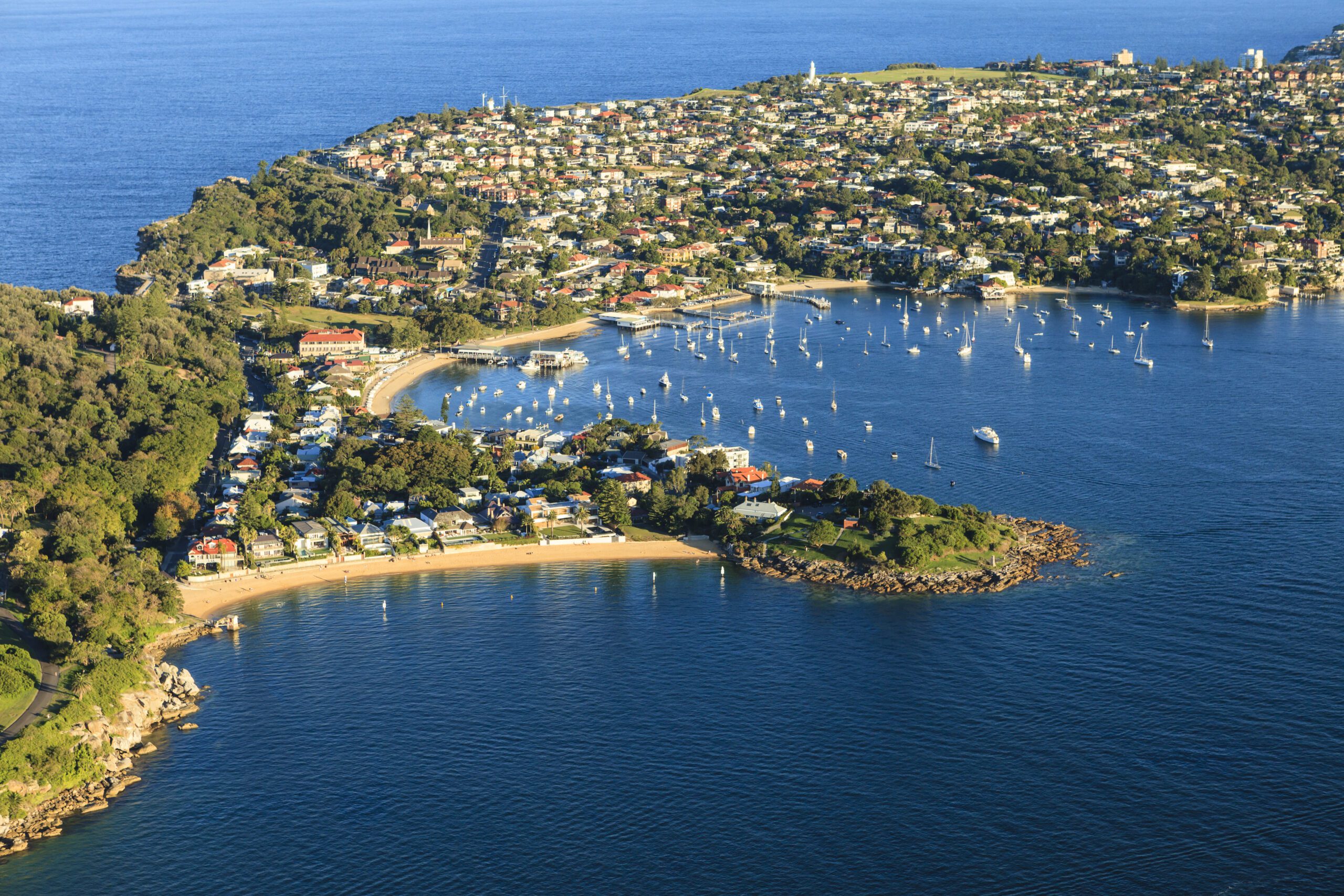 Aerial view of a coastal town with numerous houses, surrounded by lush greenery. The town encircles a bay filled with anchored boats. In the foreground is a small sandy beach and rocky shores, all bordered by the deep blue sea. The Yacht Social Club Sydney Boat Hire offers exclusive experiences here.