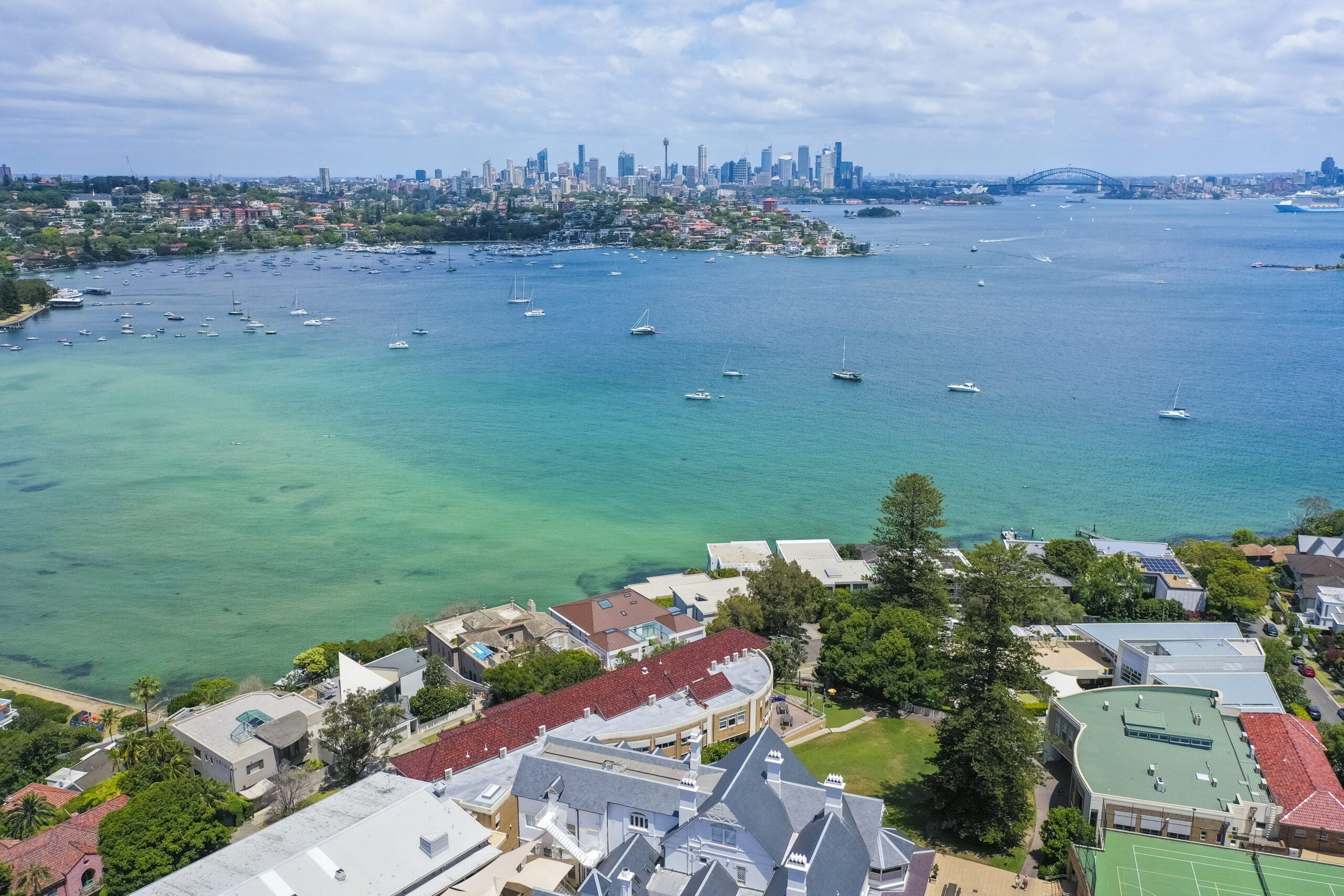 Aerial view of a picturesque coastal landscape featuring a tranquil bay with numerous sailboats. In the background, a city skyline is visible, including a prominent bridge. The foreground shows residential buildings and lush greenery, ideal for Luxury Yacht Rentals Sydney. The sky is partly cloudy.