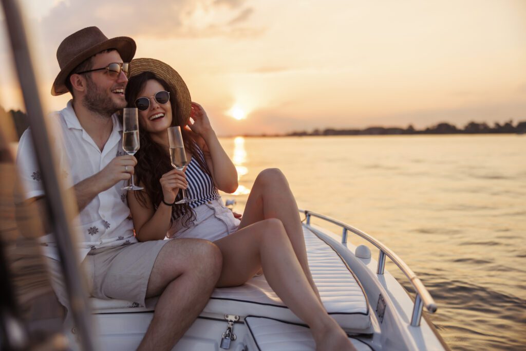 A couple in summer attire relaxes and smiles on a boat at sunset, holding glasses of champagne. They both wear hats and sunglasses, enjoying the scenic view of Sydney Harbour Boat Hire The Yacht Social Club. The golden light of the setting sun adds warmth to the scene.