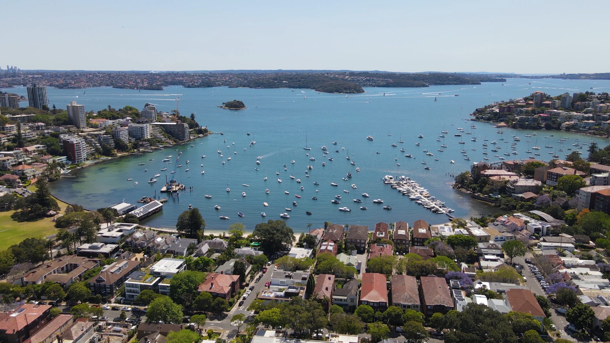 Aerial view of an urban coastal area with numerous buildings and houses near the shoreline. The harbor, perfect for Sydney Harbour Boat Hire The Yacht Social Club, is dotted with many boats and yachts. Several small islands and peninsulas are visible in the background, all under a clear, sunny sky.