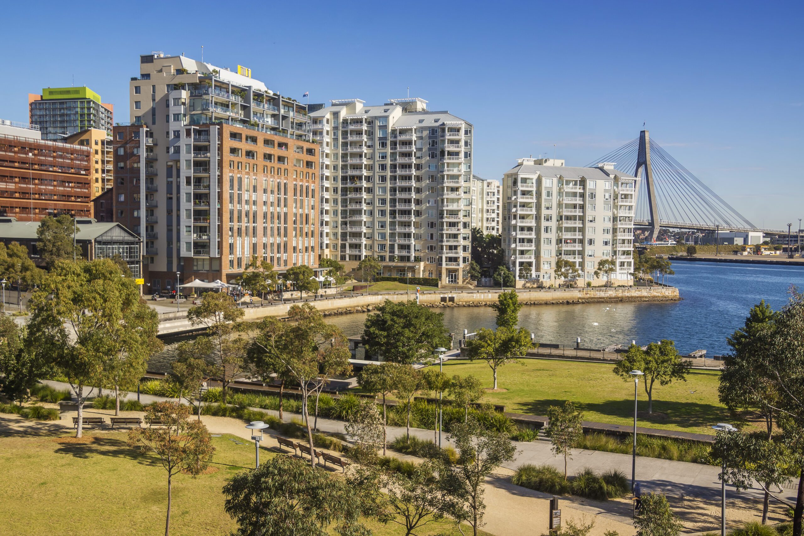 A waterfront urban park leads to a modern high-rise apartment complex. The park features walking paths, trees, and benches. In the background, a large cable-stayed bridge spans over a body of water with yachts from The Yacht Social Club gliding by, set against a clear blue sky.