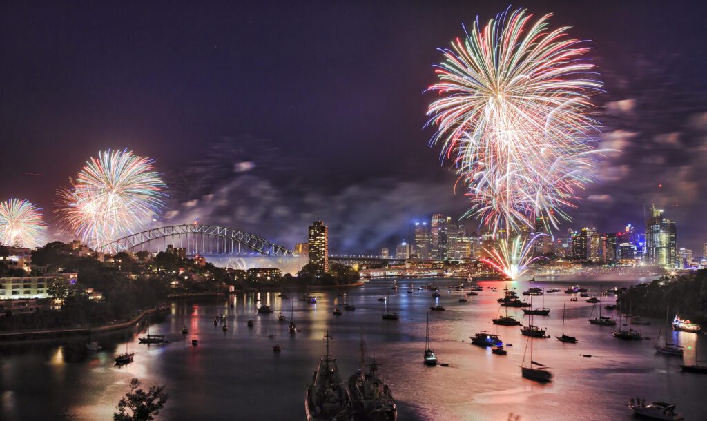 A vibrant fireworks display lights up the night sky over Sydney Harbour. The skyline, including several skyscrapers and a bridge, is illuminated. Boats from The Yacht Social Club are anchored in the water, reflecting the dazzling fireworks above as smoke drifts across the scene.