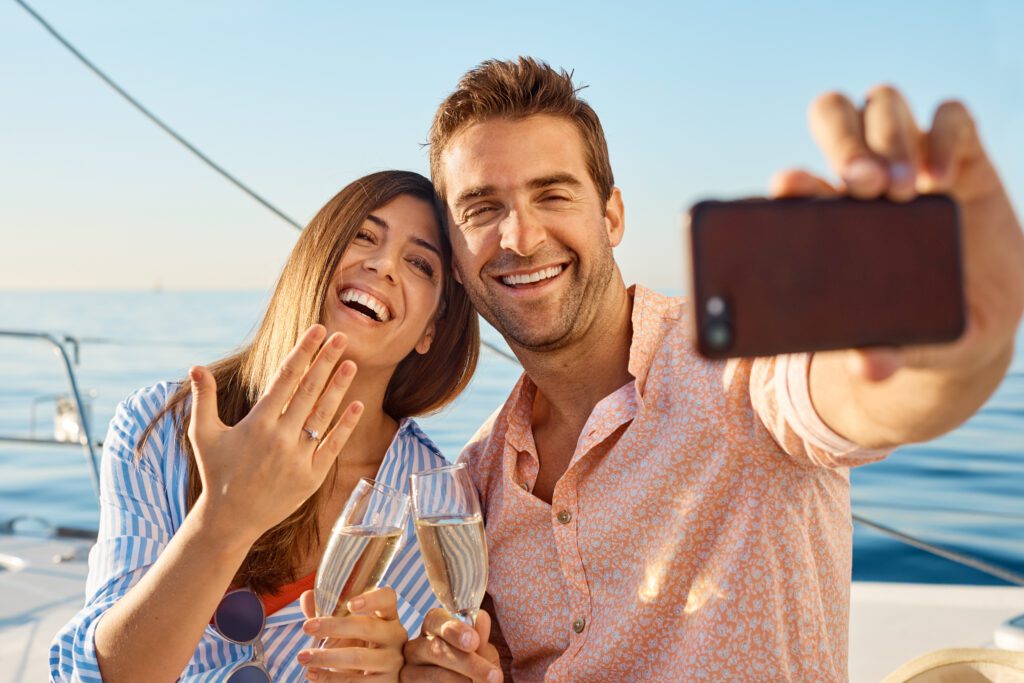 A happy couple takes a selfie on the boat from The Yacht Social Club Sydney Boat Hire. The woman shows her ring while holding a champagne glass, and the man smiles, holding the phone. Both are enjoying a sunny day by the water, with a clear blue sky and ocean in the background.