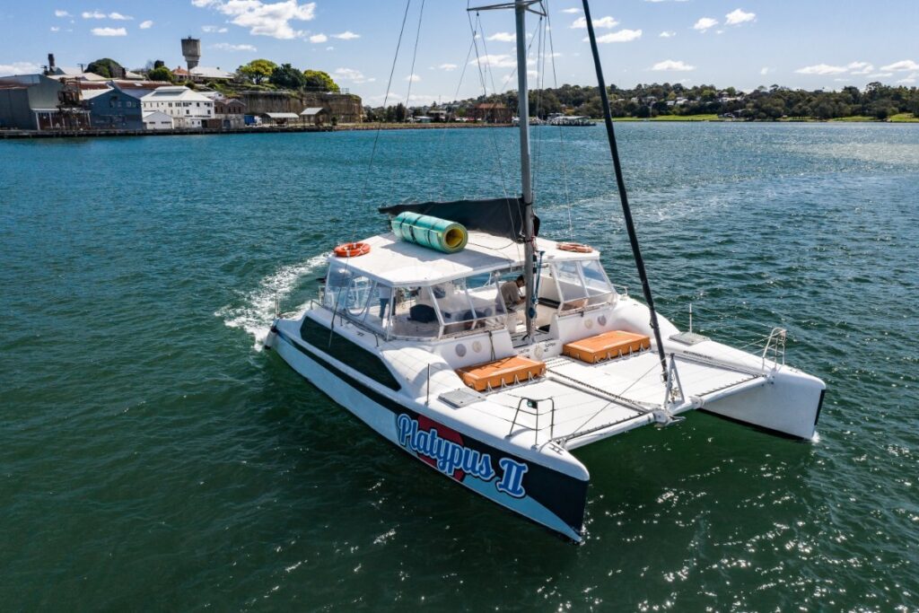 A white catamaran with "Platypus 2" written on its side sails through a calm blue body of water. It features orange cushions on the deck and a green kayak on top. In the background, buildings, trees, and a small hill sit under a blue sky with scattered clouds, making it perfect for Boat Rental and Parties Sydney The Yacht Social Club.