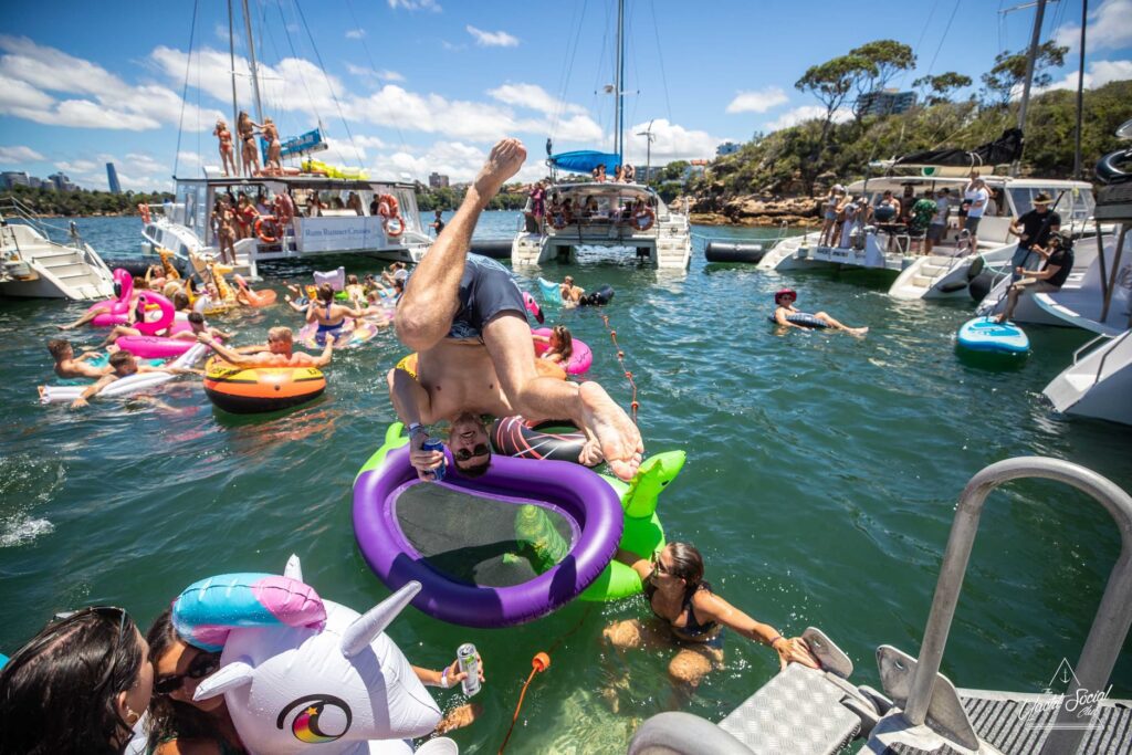 People are enjoying a sunny day on the water with multiple boats and colorful floaties. A man is enthusiastically diving headfirst off a boat onto a purple float. Others are swimming, lounging, and socializing around him, creating a lively atmosphere reminiscent of Boat Parties Sydney The Yacht Social Club.