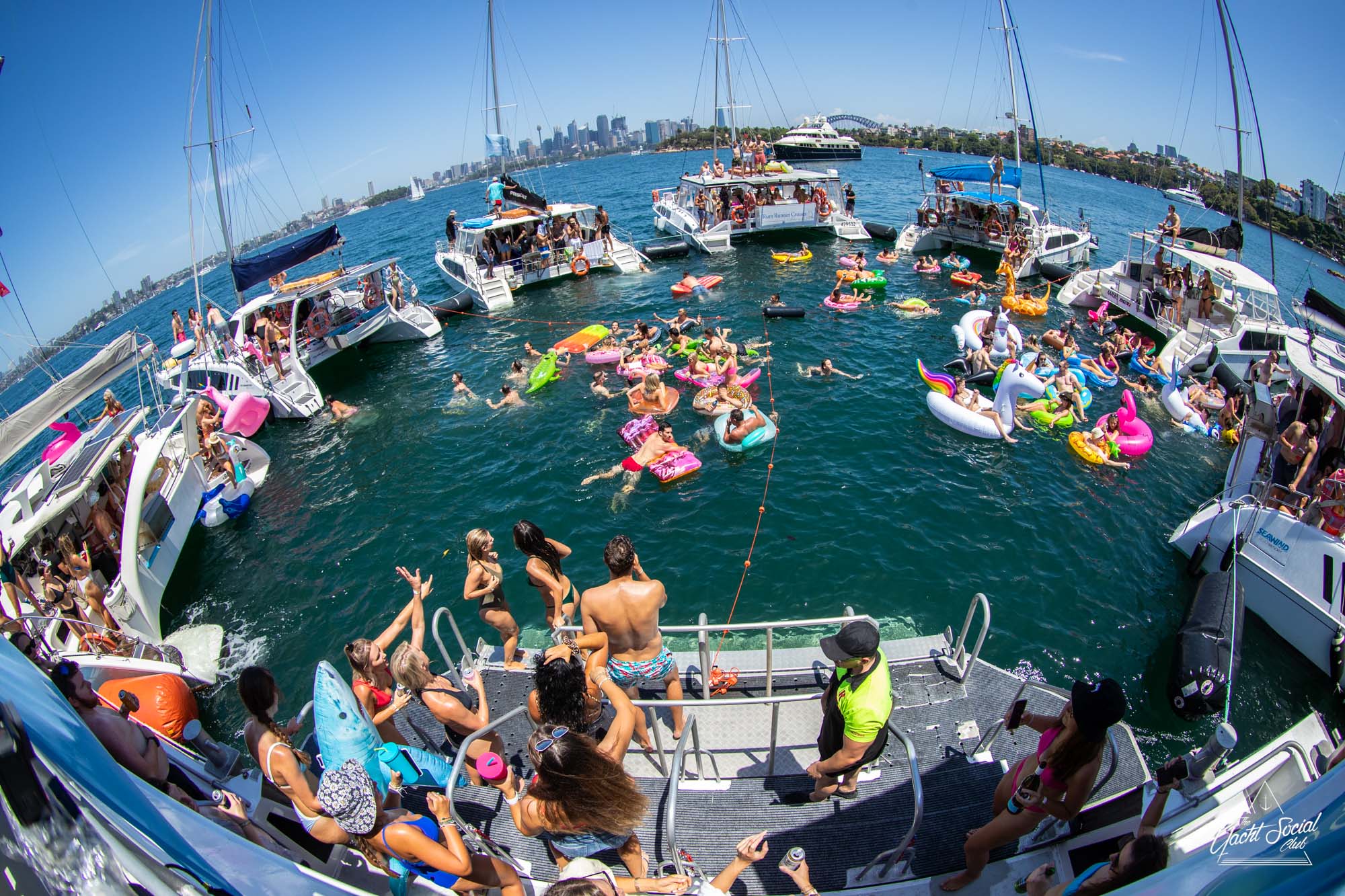 A lively boat party scene with numerous people on several interconnected boats, many of whom are on colorful inflatable floats in the water. The city skyline is visible in the background under a bright, clear sky, showcasing The Yacht Social Club Sydney Boat Hire experience.