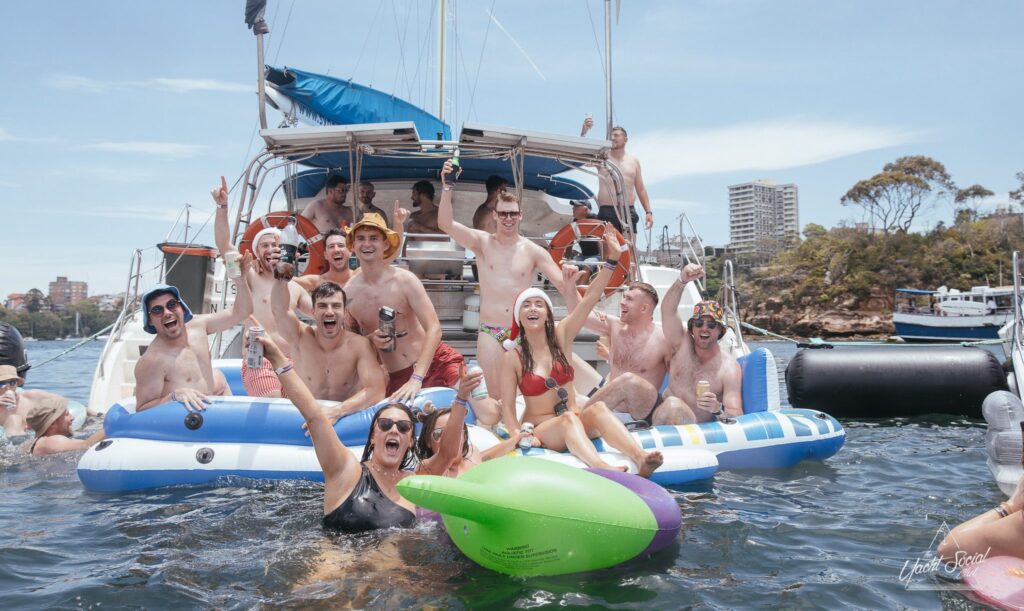 A group enjoys a sunny day on a boat and in the water. Some are seated on inflatable floats, others are standing or sitting on the boat. They are all smiling, holding drinks, and making celebratory gestures. Trees and buildings are visible in the background - a perfect scene for The Yacht Social Club Sydney Boat Hire.