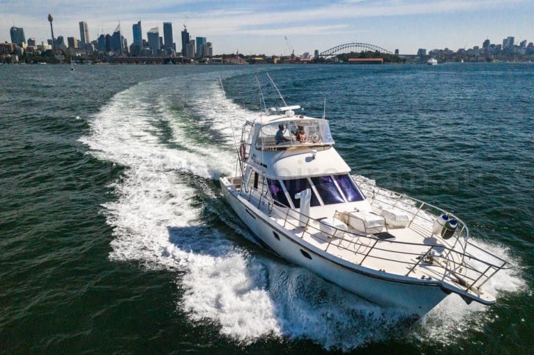 A luxury motor yacht cruises on the water with a city skyline and the Sydney Harbour Bridge in the background. The boat creates a wake behind it as it moves forward under a clear blue sky. The calm water reflects the sunlight, setting the perfect scene for Sydney Harbour Boat Hire by The Yacht Social Club.