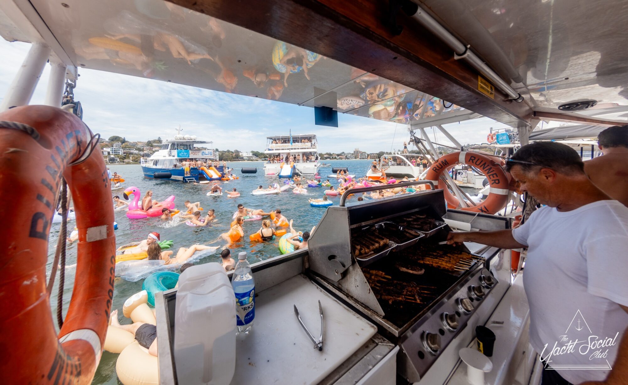 A person grills meat on a BBQ station aboard a boat. In the background, numerous people float on colorful inflatable rafts in a body of water. Other boats and a dock are also visible in the distance. The lively scene, typical of Boat Parties Sydney The Yacht Social Club, depicts a vibrant gathering.