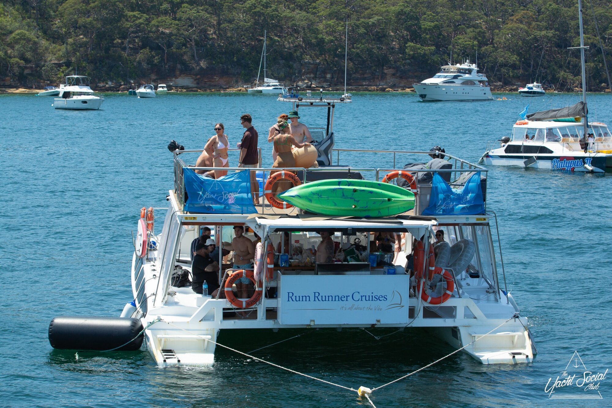 A white boat labeled "Rum Runner Cruises" is anchored on a sunny day. People are standing and sitting on the boat, some wearing swimwear. A green kayak rests atop the boat. Other boats and a lush, tree-covered shoreline are in the background, highlighting the charm of Boat Parties Sydney The Yacht Social Club.